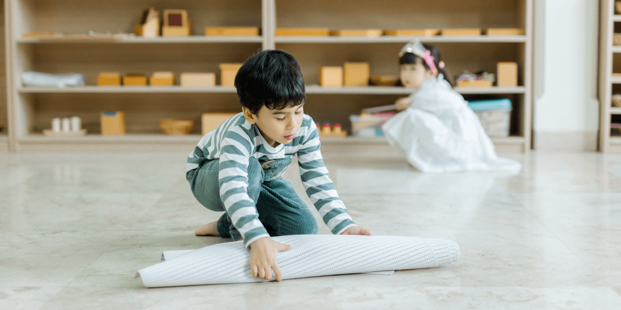 A boy rolls up his mat after use in a Montessori classroom, demonstrating respect for the learning environment and developing practical life skills.